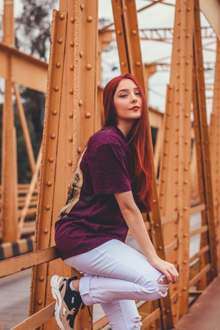 Stylish young female with long ginger hair wearing casual outfit standing on yellow metal bridge