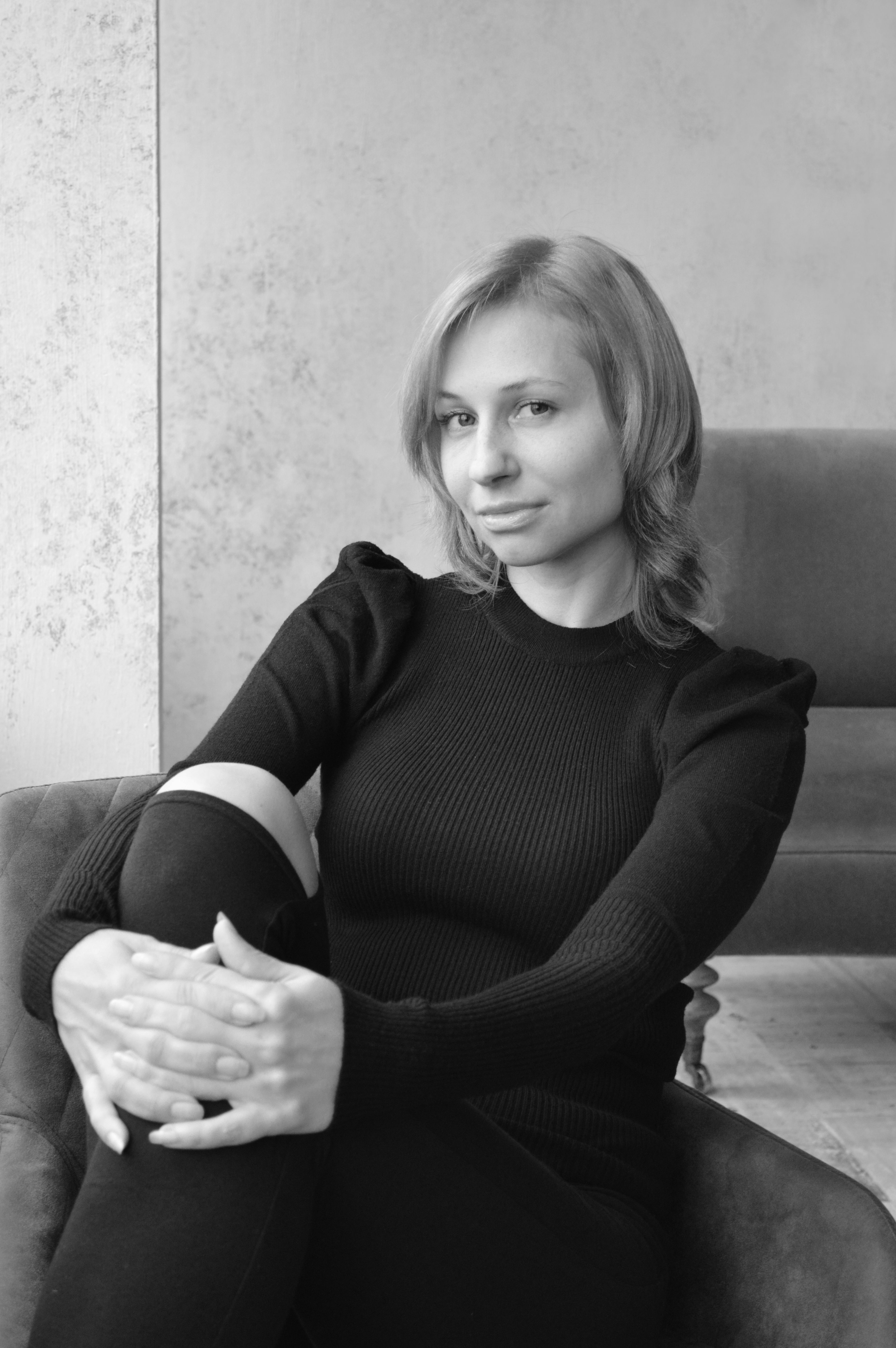 Black and white of stylish young female embracing knee and looking at camera while chilling in chair at home