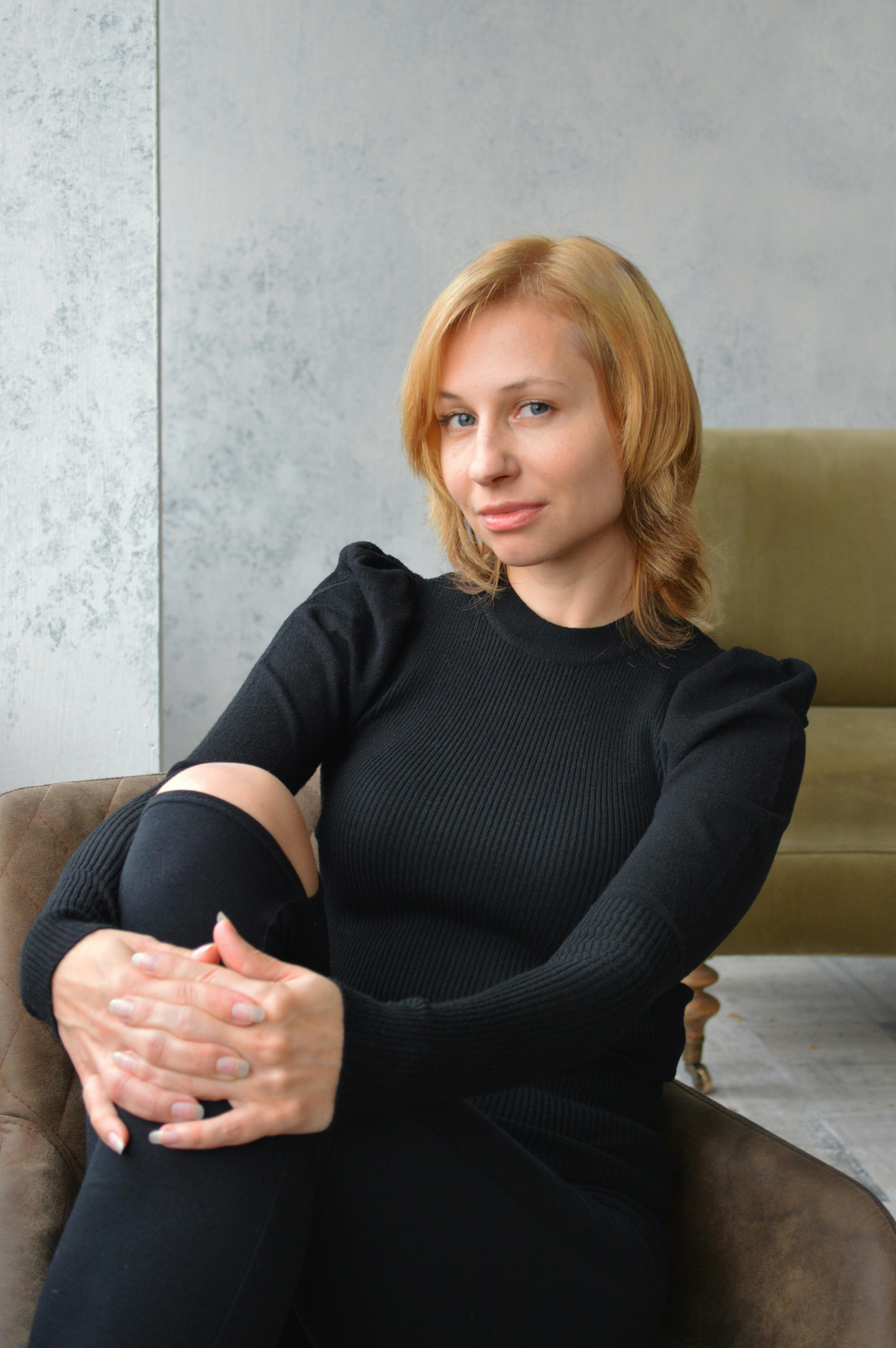 Charming coquettish young blonde female in black clothes embracing knee and looking at camera with smile while resting on soft armchair at home