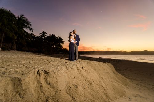 Free A couple embraces on a sandy beach at sunset with palm trees and ocean waves in the background. Stock Photo
