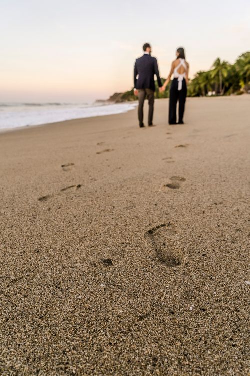 Free Couple walking hand in hand on a serene beach at sunset, leaving footprints in the sand. Stock Photo