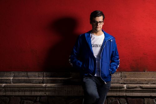 Free Man casually leaning against a vibrant red wall, wearing glasses and a blue jacket. Stock Photo
