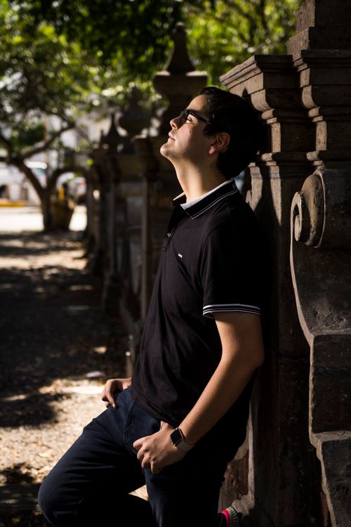 Free A man in sunglasses relaxes, leaning against a stone wall under dappled sunlight in a serene outdoor setting. Stock Photo