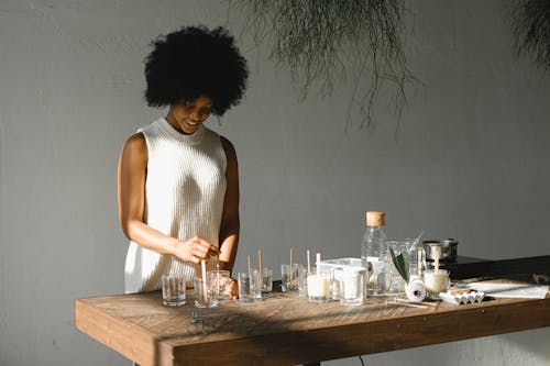 Positive African American female putting wooden wicks into candle molds while standing at wooden table with various equipment in light room