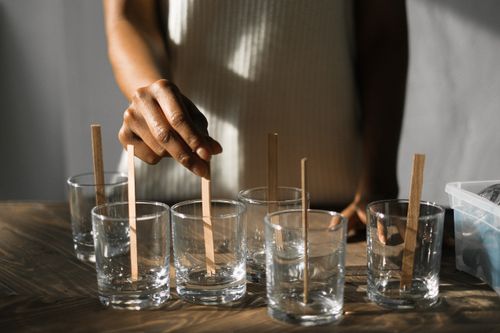 Free Unrecognizable African American female securing wooden wicks in glass molds while making aroma candles at wooden table in light studio Stock Photo