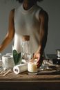 Unrecognizable African American female putting aroma candle with wooden wick on table with various equipment while working in modern studio