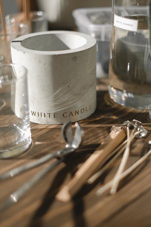 Glass jars and candle mold placed on table with steel trimmer and wooden holders for wicks in light room on blurred background