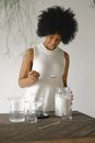 Positive African American female with spoon standing at wooden table with glass jar of wax and beakers in light studio