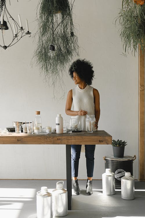 Full body of positive African American female with jar of wax pellets standing at table with various ingredients for candle making