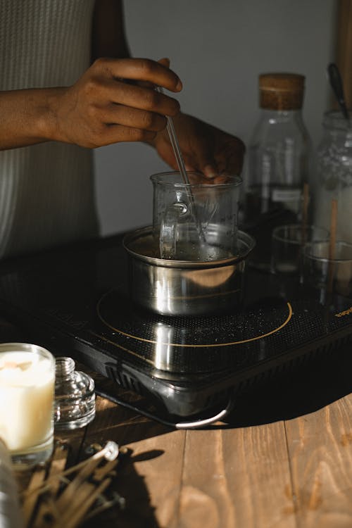 Anonymous African American female melting wax in metal pot at wooden table with cooker while making candle in light room