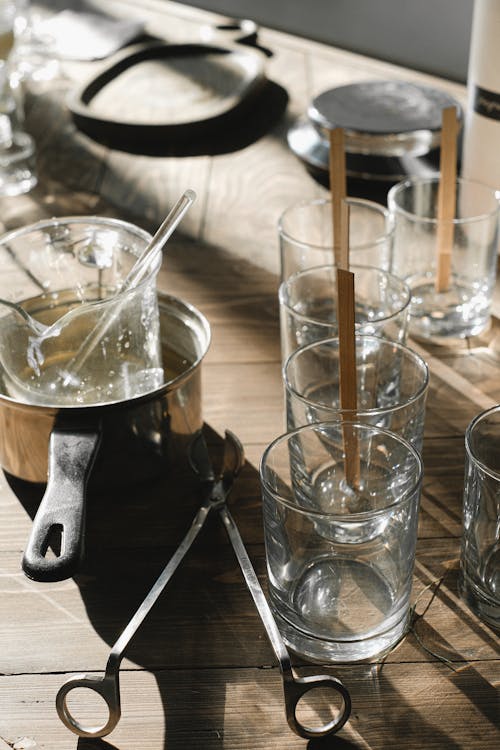 From above set of empty glass molds with wooden wicks placed on table near beaker in pot and trimmer for candle making