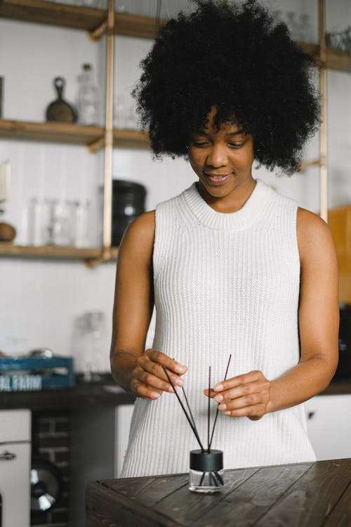 Delighted African American female standing at table with bottle with essential oil and incense sticks for aromatherapy at home