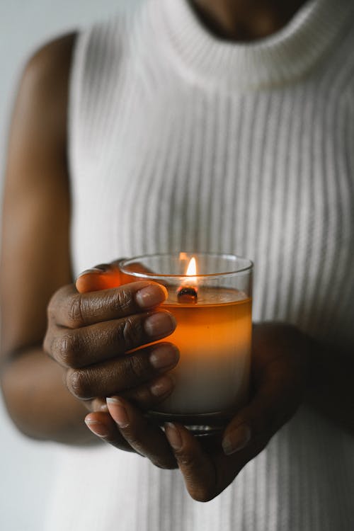 Crop anonymous black female standing with burning candle in glass for aromatherapy at home