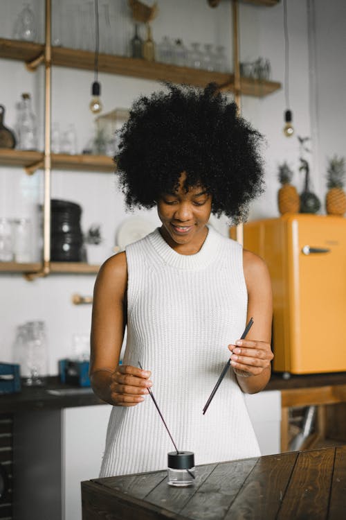 Tender smiling black female standing at table with glass bottle of essential oil and aromatic incense sticks for aromatherapy