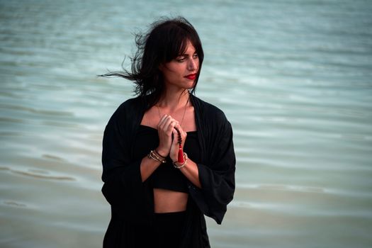 Thoughtful woman with red lips and dark hair looking away in wind weather near sea in daytime