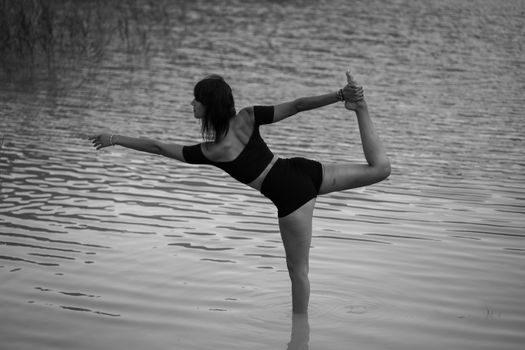 Black and white of anonymous woman in sportswear doing yoga asana Lord of the Dance in water in nature