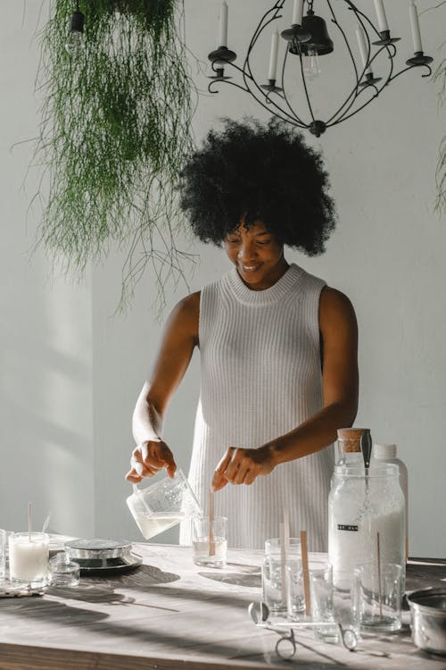 Smiling African American female standing at table and pouring liquid wax in glass while making candles for aromatherapy at table