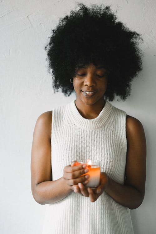 Charming African American female with curly hair standing against white wall with aromatic burning candle in glass