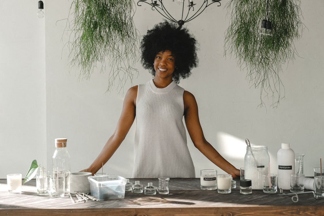 Cheerful African American craftswoman standing at table with handmade candles and assorted equipment while looking at camera