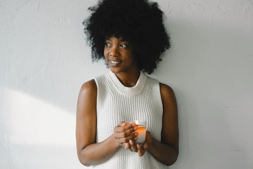 Gentle African American female with curly hair standing with aromatic burning candle in glass at home and looking away