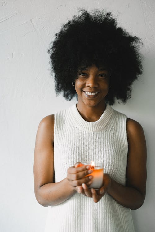Curly haired African American female standing against white wall with burning candle for aromatherapy and looking at camera