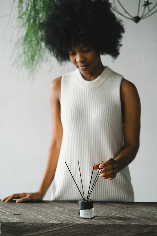 Free Peaceful African American female standing at table with incense sticks in glass bottle with essential oil and enjoying aromatherapy at home Stock Photo