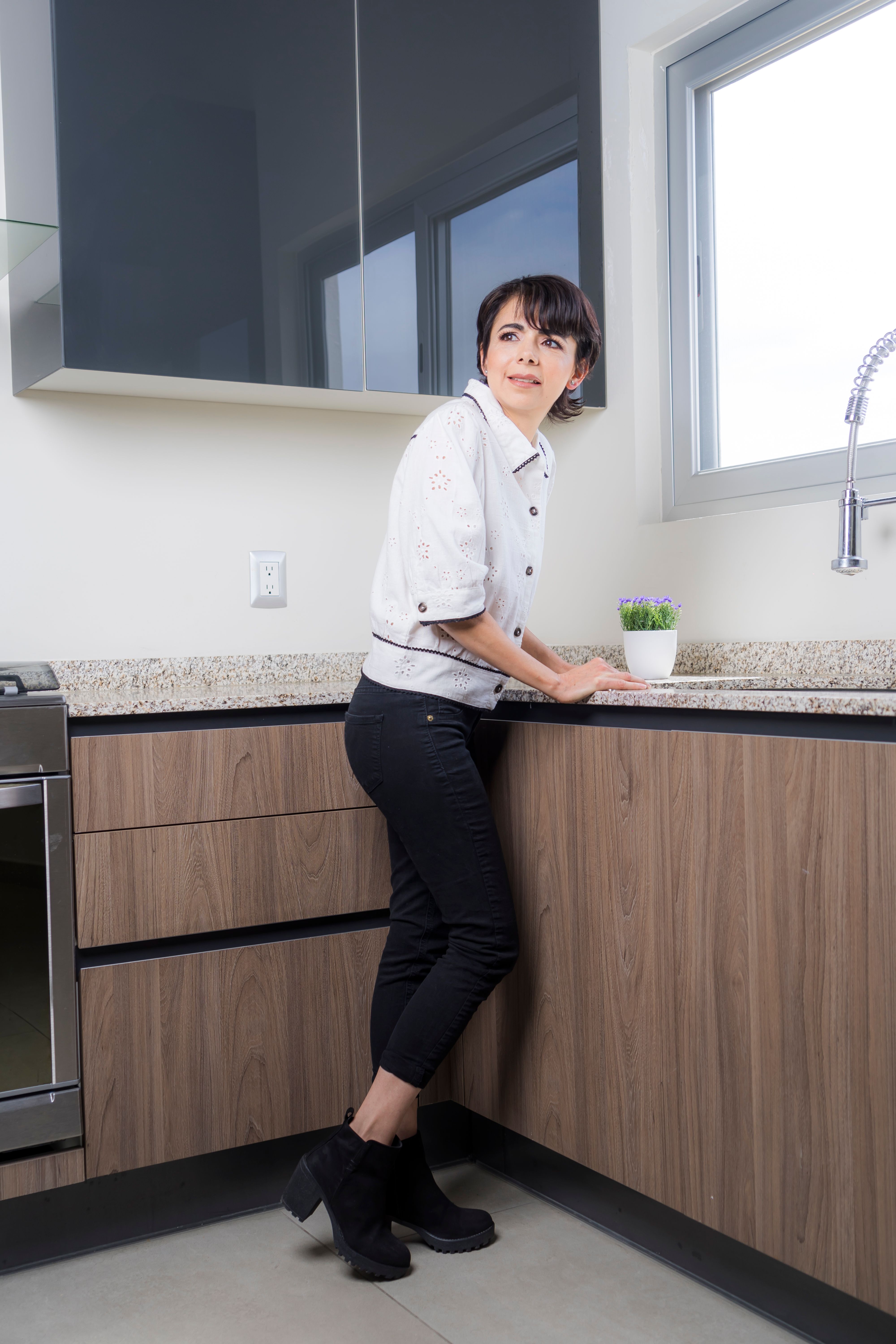 A woman stands in a modern kitchen interior smiling softly, portrays a lifestyle moment.