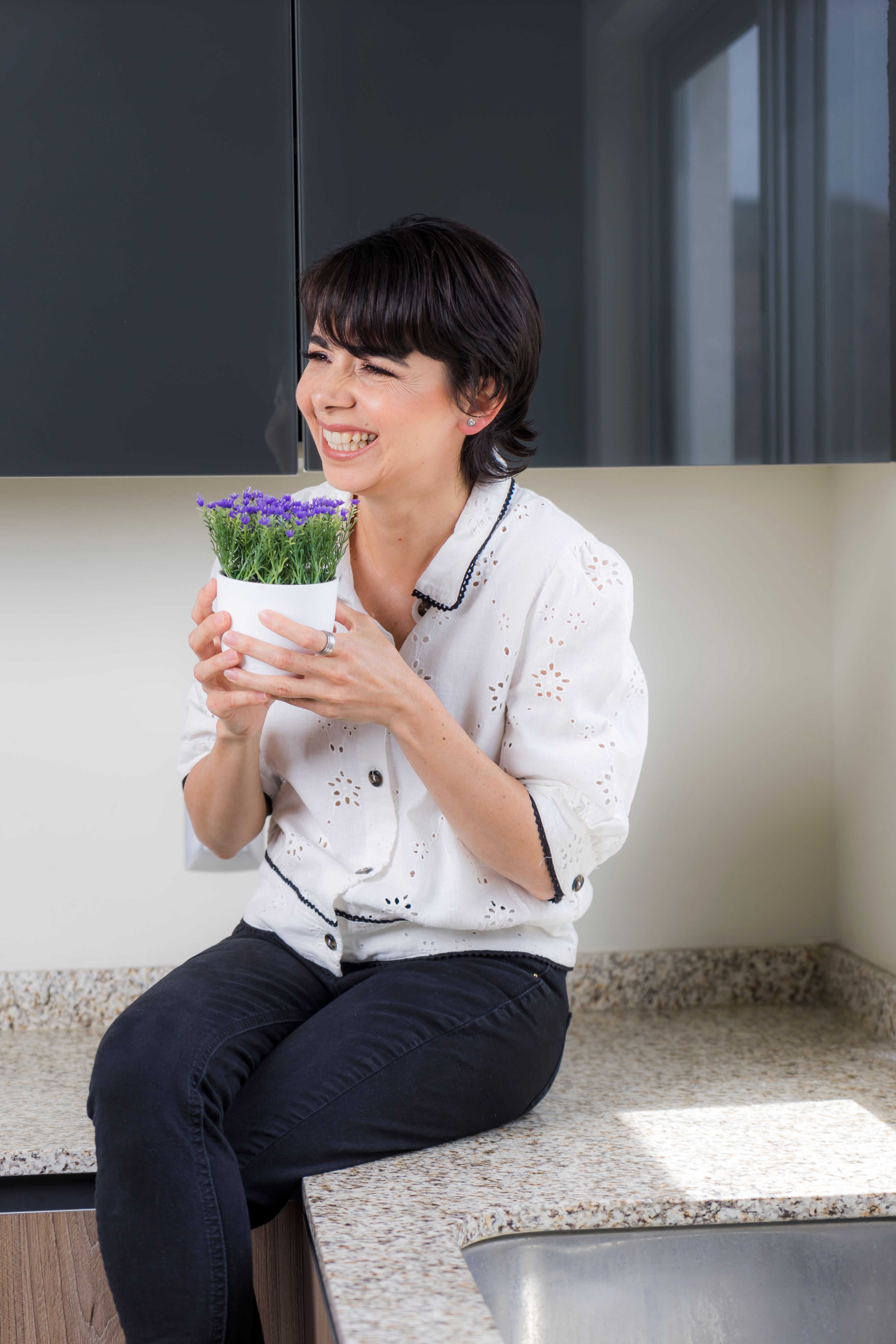 Joyful woman holding a vibrant potted plant while seated on a kitchen counter.