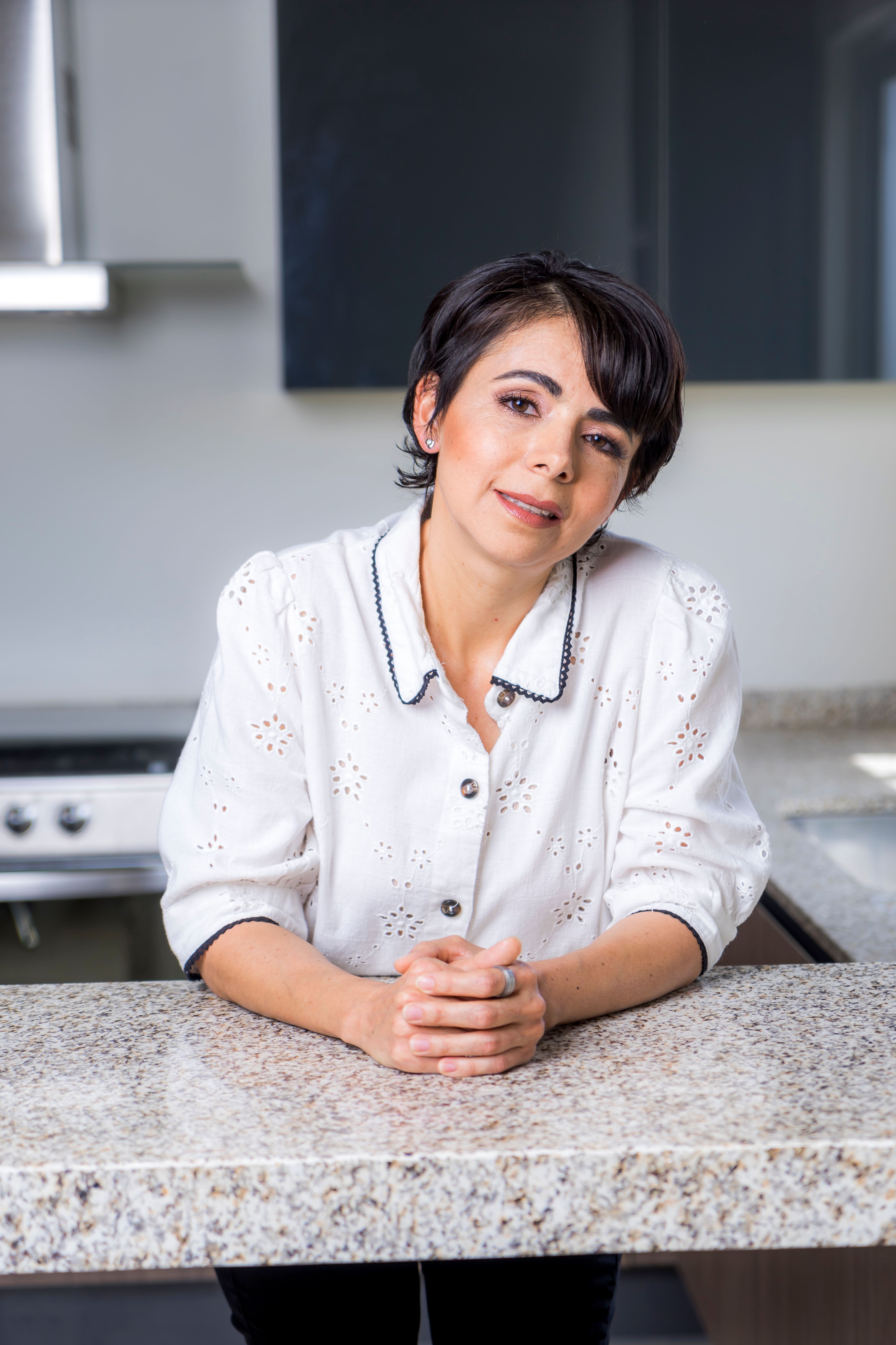 A cheerful woman in casual attire leans on a granite kitchen counter indoors.