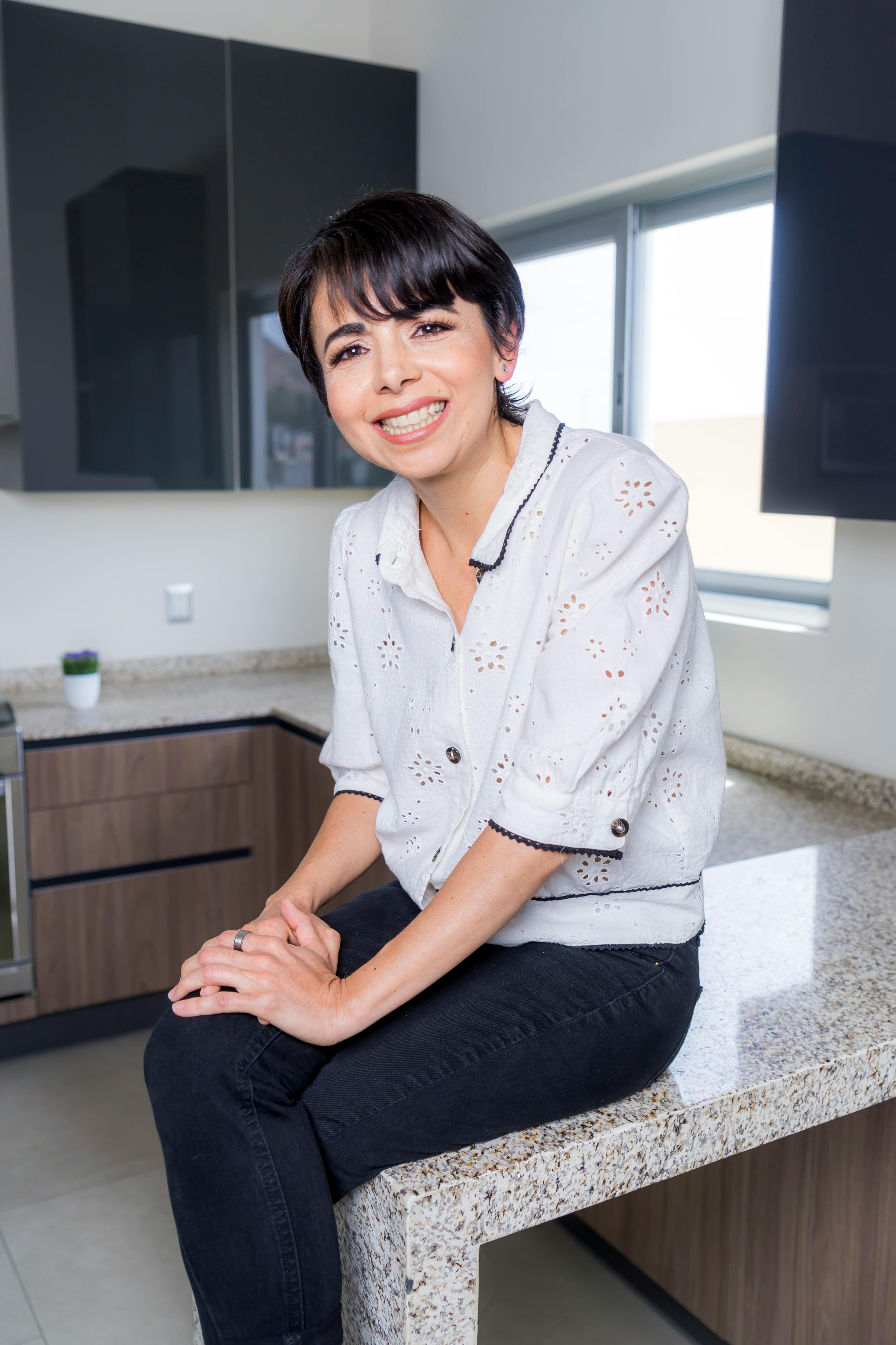 Happy Hispanic woman sitting on kitchen counter, smiling brightly in a modern kitchen.