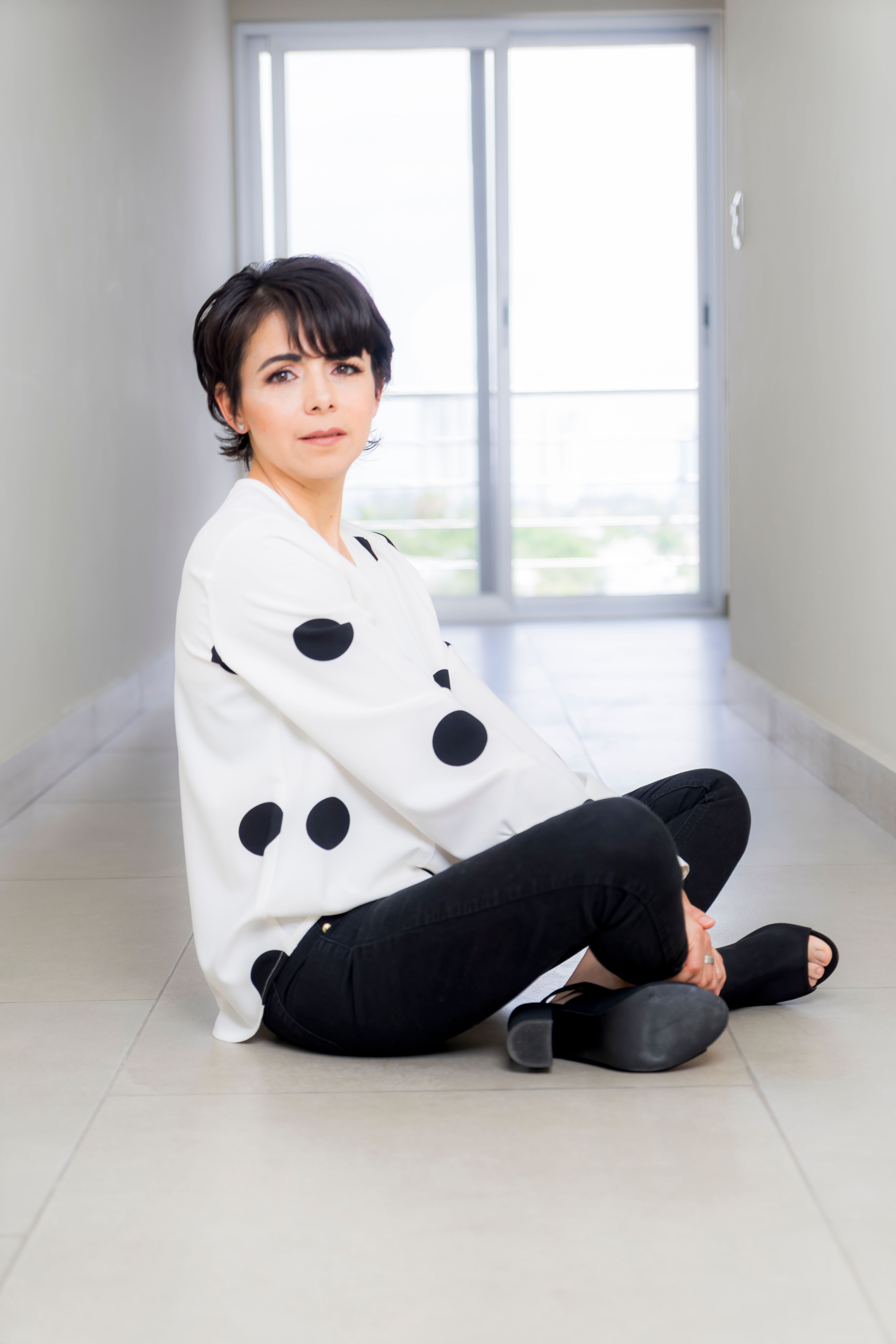 Stylish woman in black pants and white polka dot top sitting gracefully indoors.