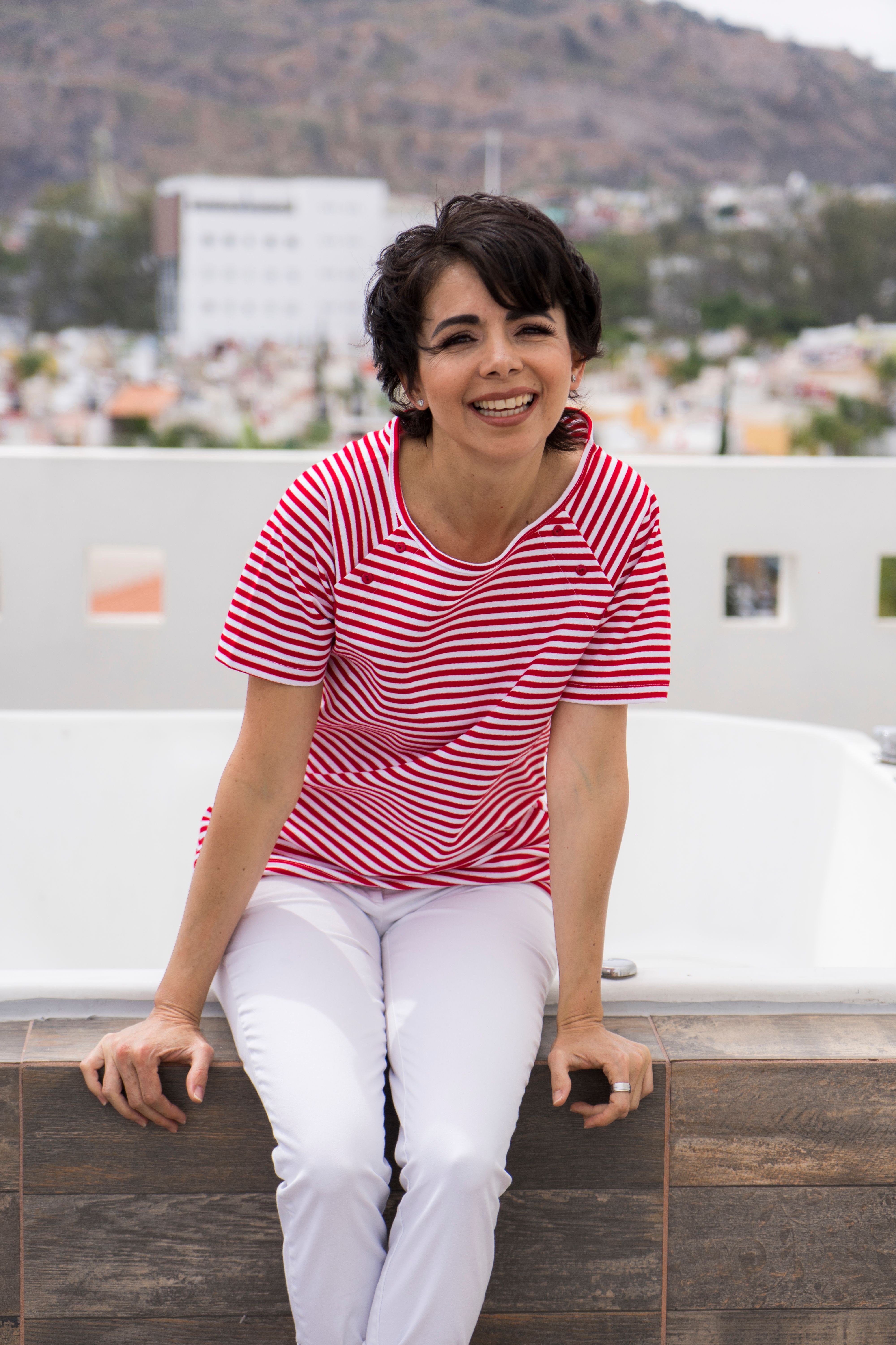 Hispanic woman with short hair in a red striped shirt sitting outdoors, smiling.