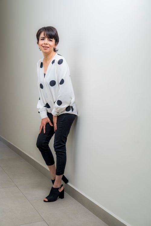 Free Stylish woman in polka dot blouse and black pants leaning against a white wall indoors. Stock Photo