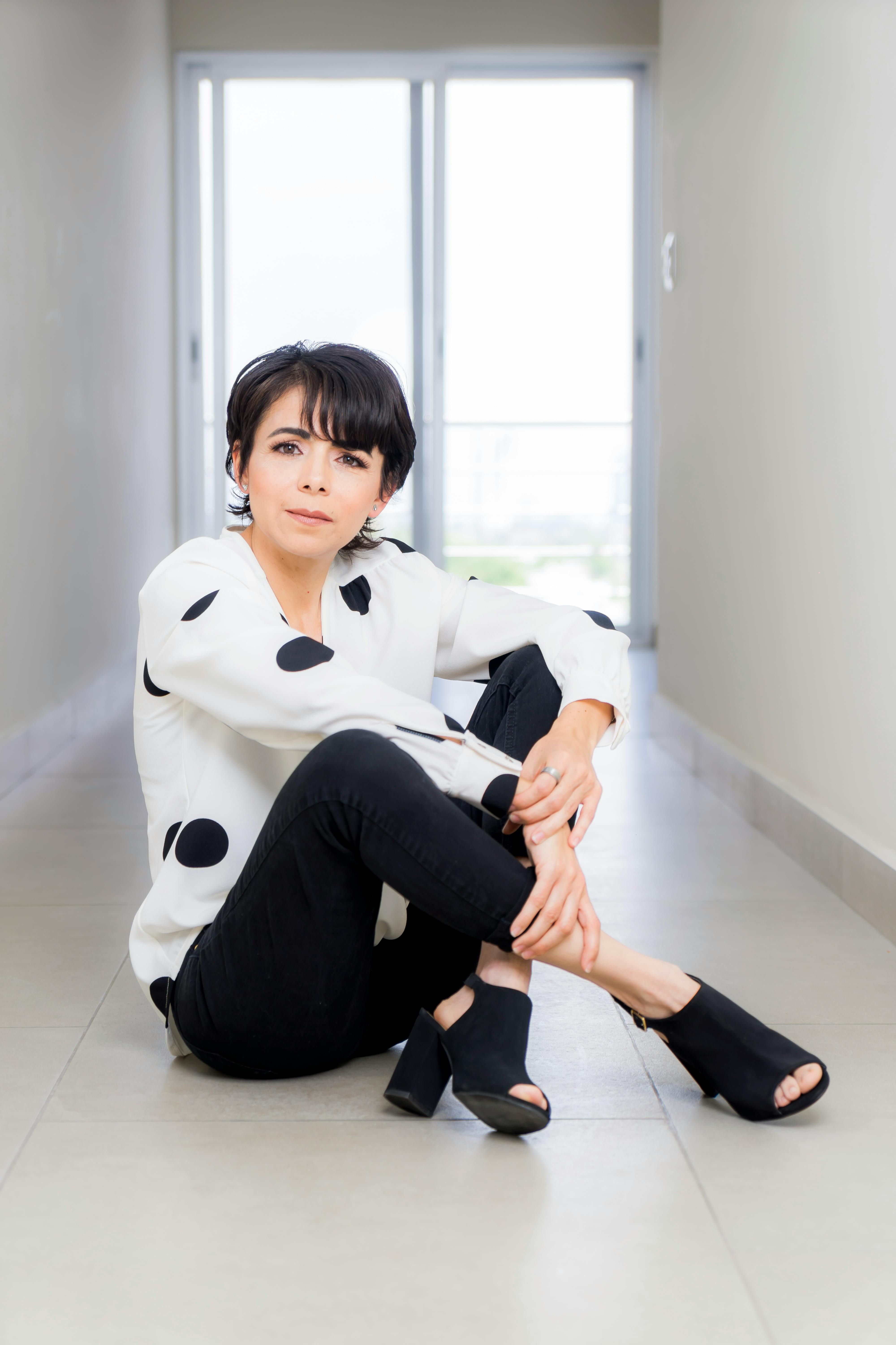 Stylish woman in black pants and polka dot blouse sitting in a bright hallway.