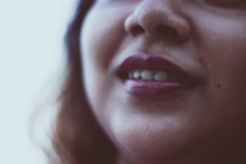 Closeup face of of unrecognizable cheerful female with dark loose hair with bright lipstick and toothy smile on blurred background in daylight