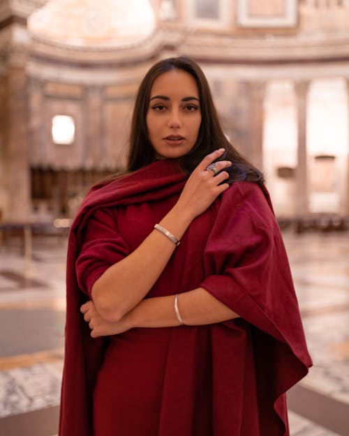 Charming young brunette in dark red loose dress raising arms in front and looking at camera while standing in grand majestic gallery