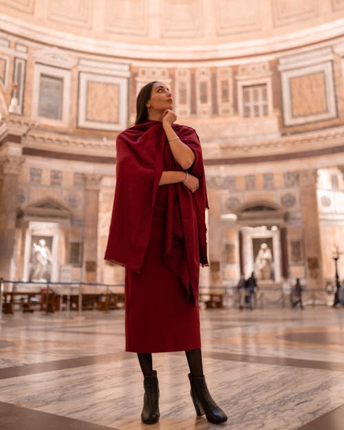Full length elegant young brunette wearing dark red dress touching chin and looking up dreamily while standing in majestic gallery