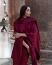 Serene young female wearing dark red fine clothes touching arm and looking away while standing near marble pillar outside majestic building