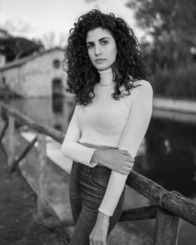 一位年轻漂亮、黑发卷曲的女子，身着黑白相间的衣服，一只手臂交叉在身前，站在宁静乡村河岸的木栅栏旁