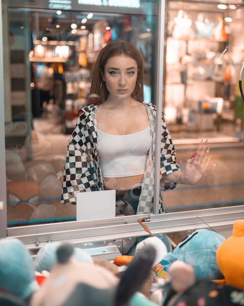 Calm wistful female in stylish wear standing behind glass prize machine and looking at camera while spending time in shopping center