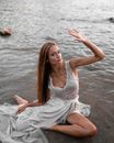 From above full body gorgeous young female wearing fancy maxi dress resting on wet seacoast with arm raised and looking at camera