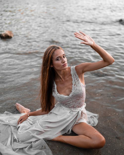 Free From above full body gorgeous young female wearing fancy maxi dress resting on wet seacoast with arm raised and looking at camera Stock Photo