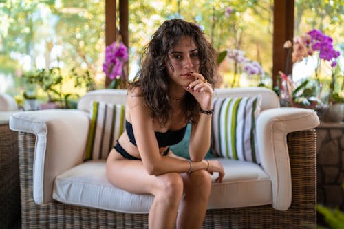 Free Young slim female with messy hair and in bikini looking at camera and relaxing on modern sofa on blurred background of interior of home Stock Photo