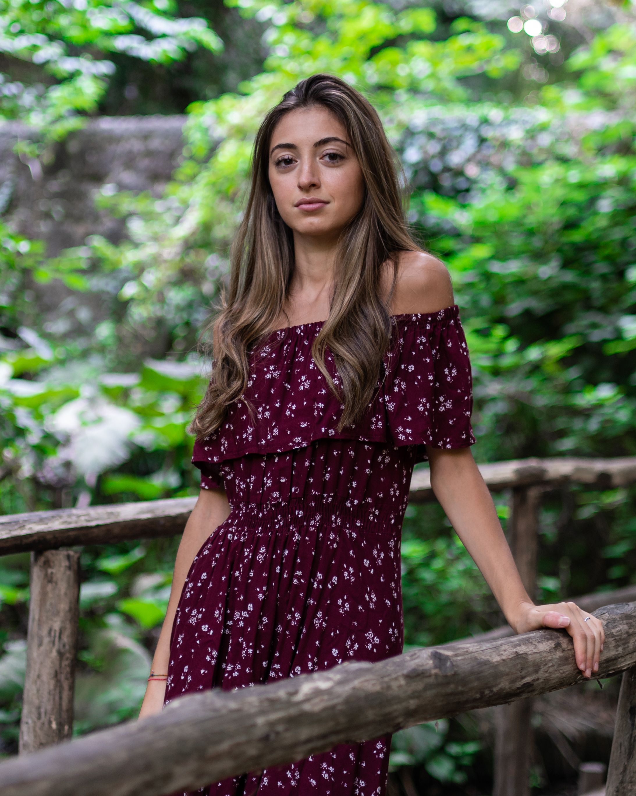 Young woman with long wavy hair in stylish red dress standing on wooden bridge in green forest and looking at camera