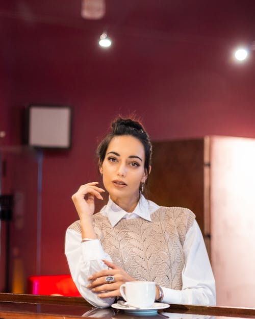 Young slim female in casual clothes leaning on bar rack with cup of hot beverage and looking at camera