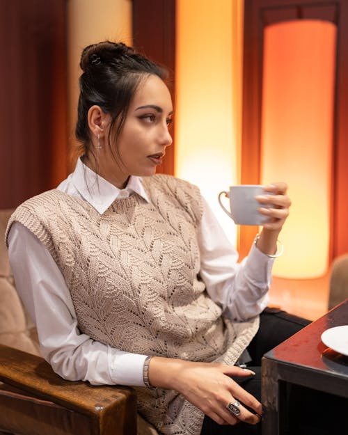 Young slim female in casual outfit and earrings sitting in armchair near table and holding cup of hot coffee