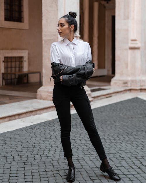 Full body of confident female in stylish outfit standing near entrance of old building with crossed arms and looking away