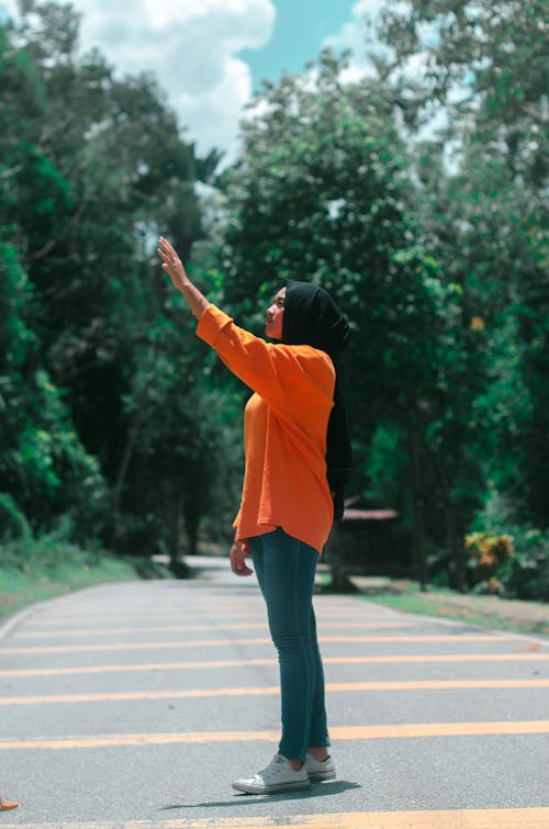 Woman in Orange Long Sleeve Shirt and Denim Pants Standing on the Road