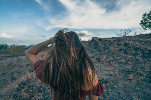 Back view of anonymous female touching hair on land against hillside under blue cloudy sky in daylight