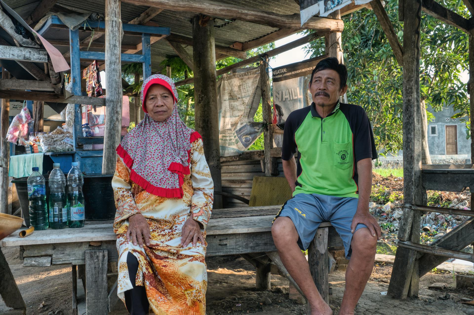 Calm elderly Asian native female in casual clothes and hijab sitting on wooden bench near middle aged man in traditional village